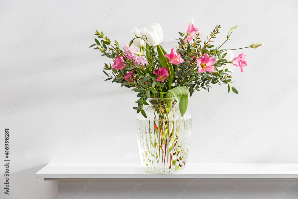 Fototapeta premium A minimalistic bouquet of white tulips, pink eustoma, hyacinth, eucalyptus in a fluted glass vase on a white panel of an artificial fireplace.