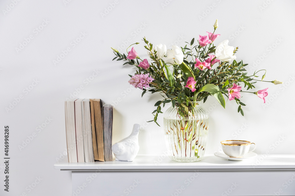 A minimalistic bouquet of white tulips, pink eustoma, hyacinth, eucalyptus in a fluted glass vase on a white panel of an artificial fireplace.