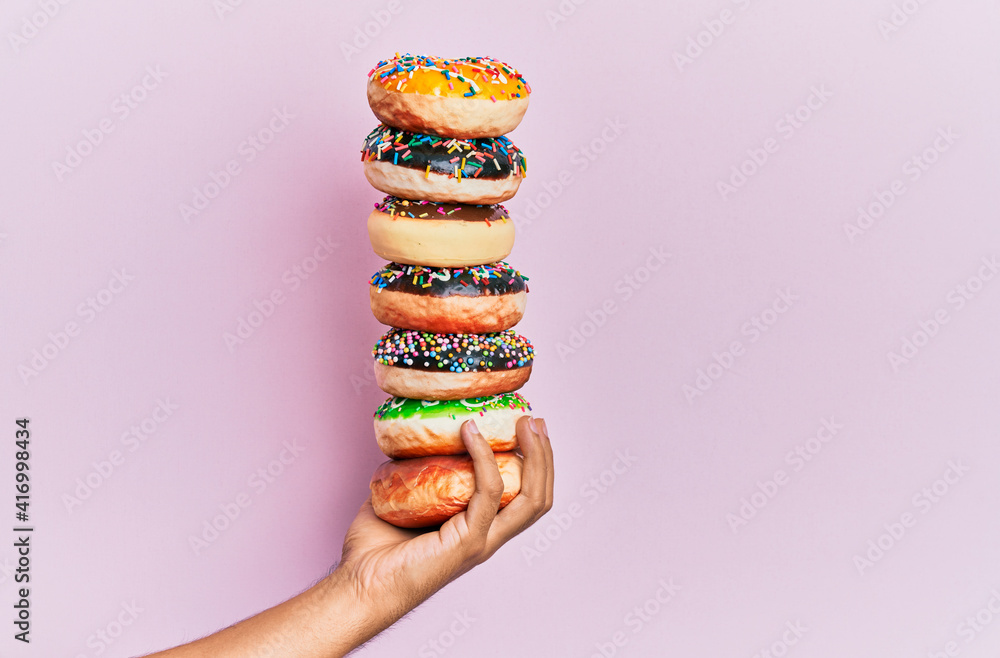 © Krakenimages.com - Hand of hispanic man holding tower of donuts over isolated pink background.