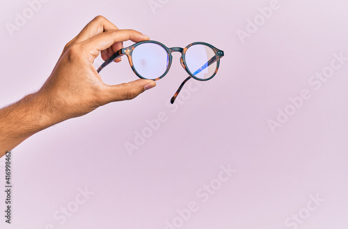 Hand of hispanic man holding glasses over isolated pink background.