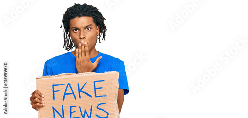 Young african american man holding fake news banner covering mouth with hand, shocked and afraid for mistake. surprised expression © Krakenimages.com
