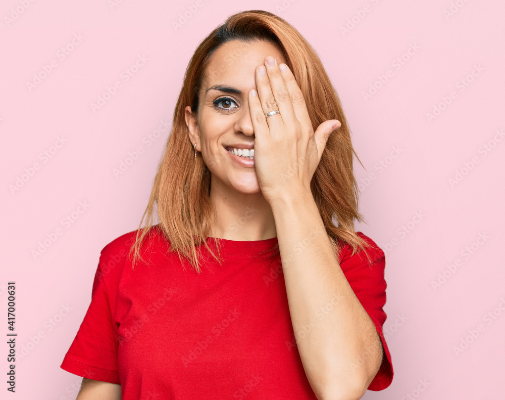 Hispanic young woman wearing casual red t shirt covering one eye with hand, confident smile on face and surprise emotion. Hispanic young woman wearing casual red t shirt covering one eye with hand, confident smile on face and surprise emotion.