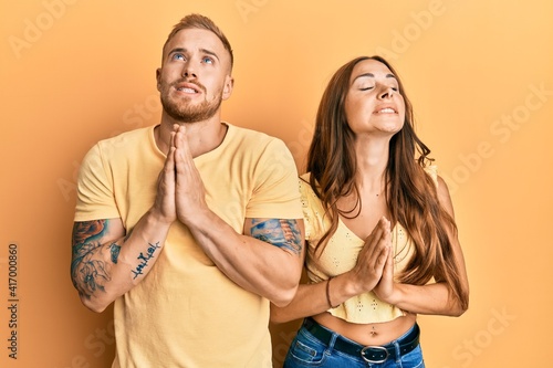 Young couple of girlfriend and boyfriend hugging and standing together begging and praying with hands together with hope expression on face very emotional and worried. begging.