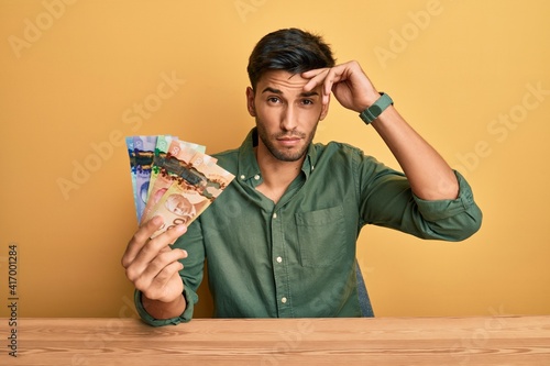 Young handsome man holding canadian dollars worried and stressed about a problem with hand on forehead, nervous and anxious for crisis