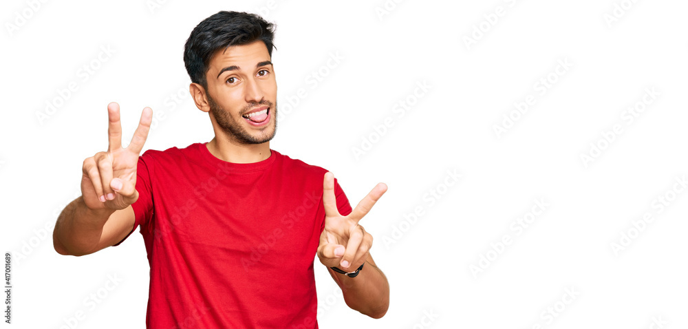 Young handsome man wearing casual red tshirt smiling with tongue out showing fingers of both hands doing victory sign. number two. Young handsome man wearing casual red tshirt smiling with tongue out showing fingers of both hands doing victory sign. number two.