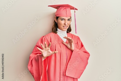 Young caucasian woman wearing graduation cap and ceremony robe disgusted expression, displeased and fearful doing disgust face because aversion reaction. with hands raised