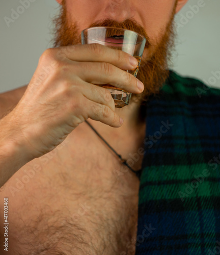 Irish bearded man in traditional green kilt celebrates St. Patrick's Day with scotch drink glass in hand. Scottish guy drinks whiskey in pub portrait.17 March cultural grand parade celebration Ireland