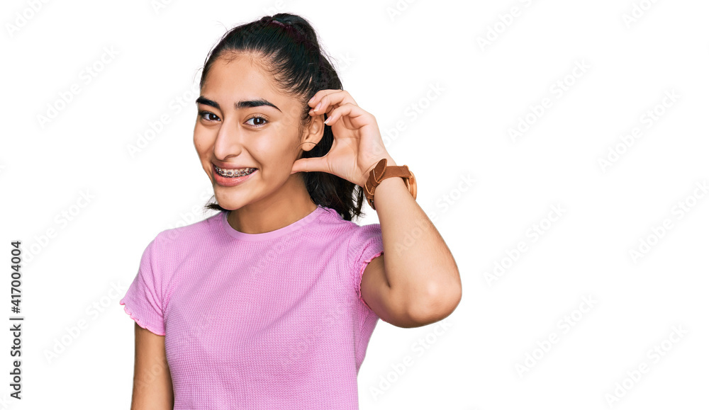 Hispanic teenager girl with dental braces wearing casual clothes smiling with hand over ear listening an hearing to rumor or gossip. deafness concept. Hispanic teenager girl with dental braces wearing casual clothes smiling with hand over ear listening an hearing to rumor or gossip. deafness concept.