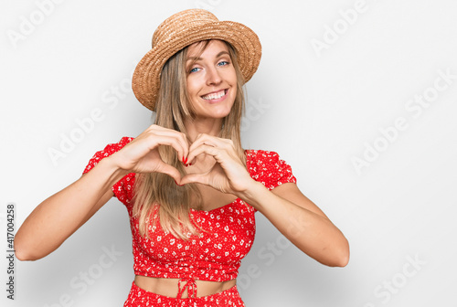 Beautiful caucasian woman wearing summer hat smiling in love doing heart symbol shape with hands. romantic concept.
