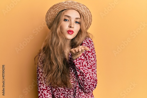 Beautiful blonde caucasian woman wearing summer hat looking at the camera blowing a kiss with hand on air being lovely and sexy. love expression.