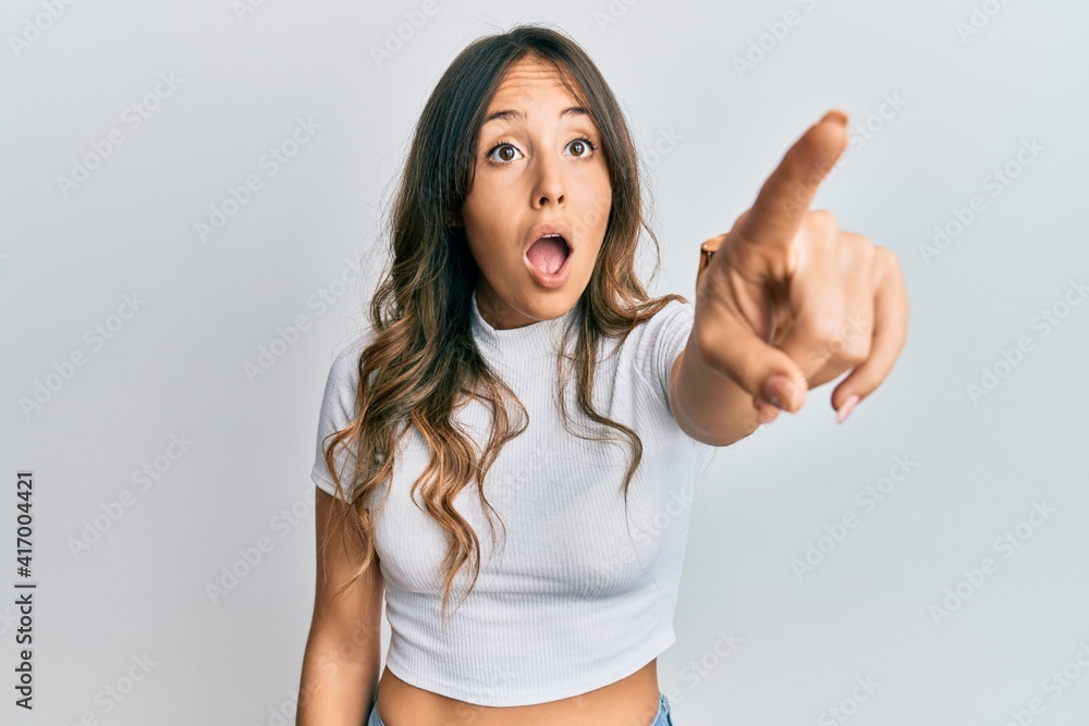 Young brunette woman wearing casual white t shirt pointing with finger surprised ahead, open mouth amazed expression, something on the front Young brunette woman wearing casual white t shirt pointing with finger surprised ahead, open mouth amazed expression, something on the front