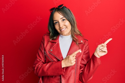 Young brunette woman wearing red leather jacket smiling and looking at the camera pointing with two hands and fingers to the side.