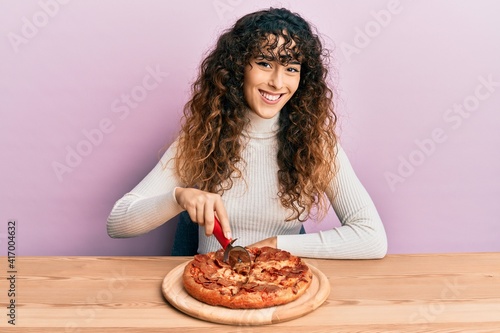 Young hispanic girl cutting tasty pepperoni pizza looking positive and happy standing and smiling with a confident smile showing teeth