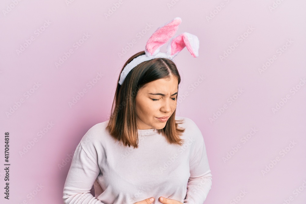 Young beautiful woman wearing cute easter bunny ears with hand on stomach because indigestion, painful illness feeling unwell. ache concept. Young beautiful woman wearing cute easter bunny ears with hand on stomach because indigestion, painful illness feeling unwell. ache concept.