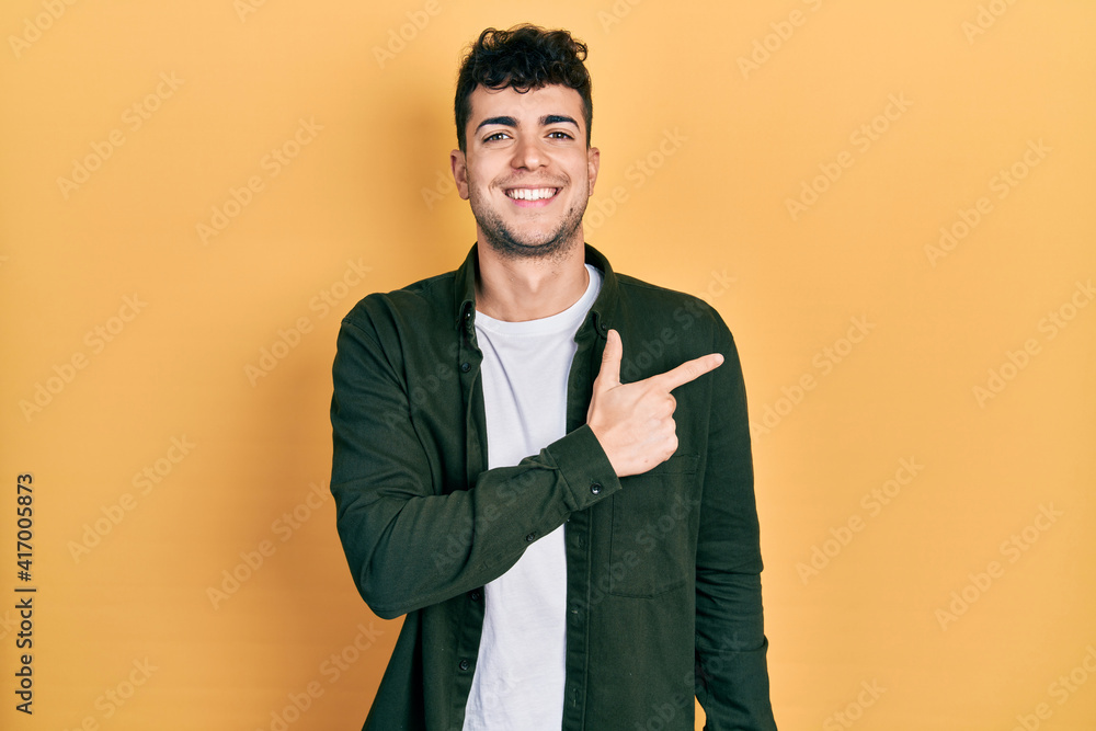 Young hispanic man wearing casual clothes cheerful with a smile of face pointing with hand and finger up to the side with happy and natural expression on face Young hispanic man wearing casual clothes cheerful with a smile of face pointing with hand and finger up to the side with happy and natural expression on face