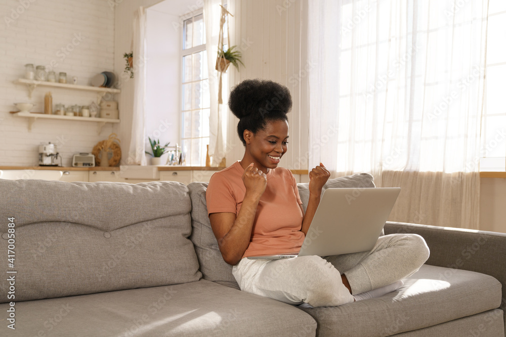 Excited millennial African woman sitting on couch at home, get pleasant online message on laptop, receive unexpected good news or email, feel euphoric. Black young girl triumph win lottery. Excited millennial African woman sitting on couch at home, get pleasant online message on laptop, receive unexpected good news or email, feel euphoric. Black young girl triumph win lottery.