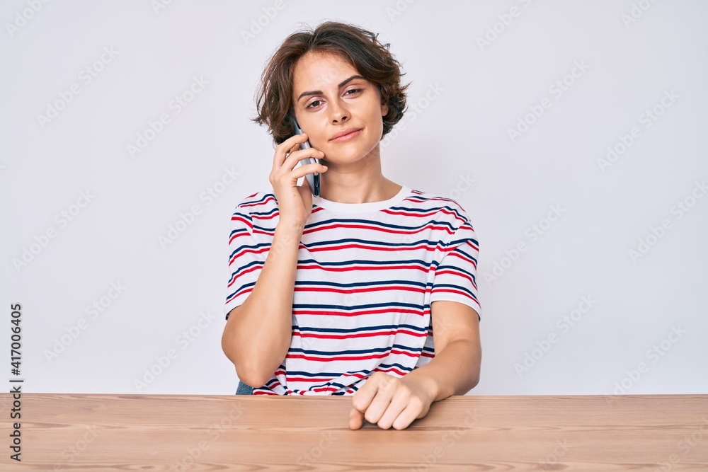 Young hispanic woman talking on the smartphone sitting on the table thinking attitude and sober expression looking self confident Young hispanic woman talking on the smartphone sitting on the table thinking attitude and sober expression looking self confident