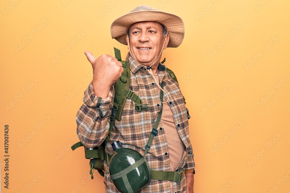 Fototapeta premium Senior man with grey hair wearing hiker backpack and water canteen pointing thumb up to the side smiling happy with open mouth