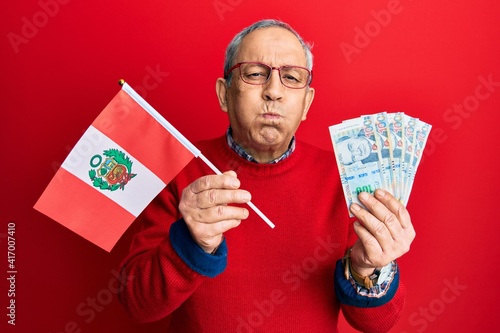 Handsome senior man with grey hair holding peru flag and peruvian sol banknotes puffing cheeks with funny face. mouth inflated with air, catching air.