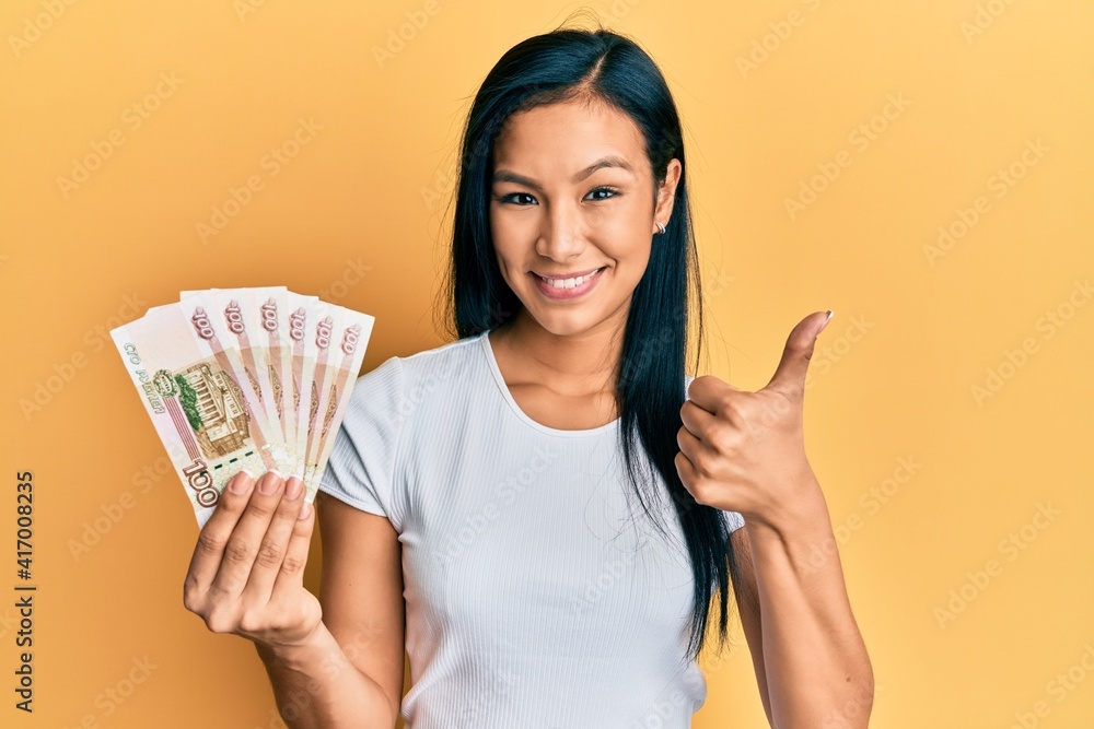 Beautiful hispanic woman holding 100 russian ruble banknotes smiling happy and positive, thumb up doing excellent and approval sign Beautiful hispanic woman holding 100 russian ruble banknotes smiling happy and positive, thumb up doing excellent and approval sign