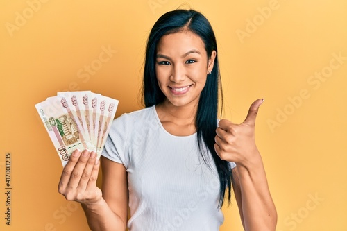 Beautiful hispanic woman holding 100 russian ruble banknotes smiling happy and positive, thumb up doing excellent and approval sign