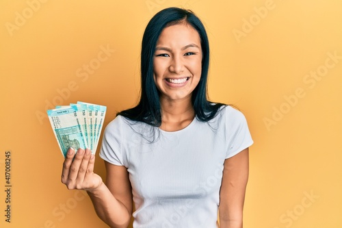 Beautiful hispanic woman holding 50 indian rupee banknotes looking positive and happy standing and smiling with a confident smile showing teeth