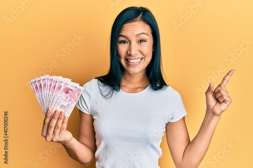Beautiful hispanic woman holding 10 turkish lira banknotes smiling happy pointing with hand and finger to the side