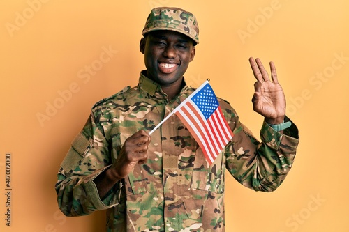 Young african american man wearing army uniform holding american flag doing ok sign with fingers  smiling friendly gesturing excellent symbol