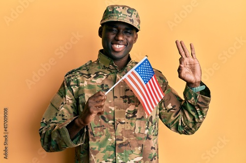 Young african american man wearing army uniform holding american flag doing ok sign with fingers, smiling friendly gesturing excellent symbol