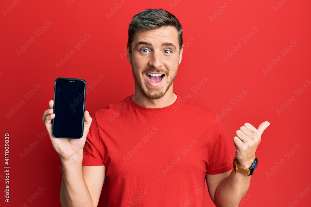 Handsome caucasian man holding smartphone showing screen pointing thumb up to the side smiling happy with open mouth Handsome caucasian man holding smartphone showing screen pointing thumb up to the side smiling happy with open mouth