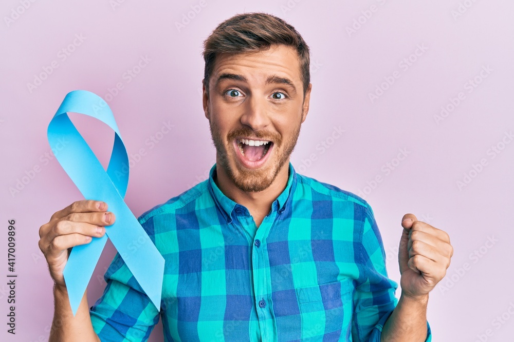 Handsome caucasian man holding blue ribbon screaming proud, celebrating victory and success very excited with raised arms Handsome caucasian man holding blue ribbon screaming proud, celebrating victory and success very excited with raised arms