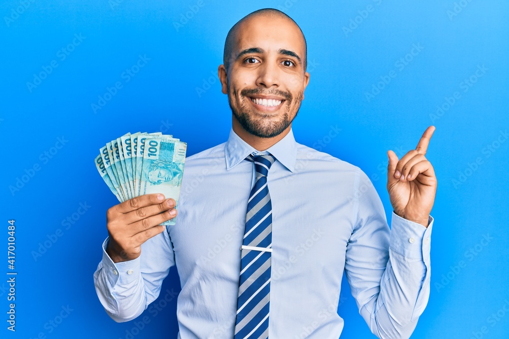 Hispanic adult man holding 100 brazilian real banknotes smiling happy pointing with hand and finger to the side Hispanic adult man holding 100 brazilian real banknotes smiling happy pointing with hand and finger to the side