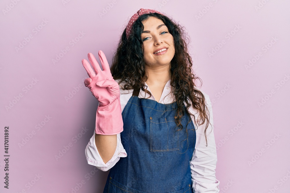 Young brunette woman with curly hair wearing cleaner apron and gloves showing and pointing up with fingers number three while smiling confident and happy. Young brunette woman with curly hair wearing cleaner apron and gloves showing and pointing up with fingers number three while smiling confident and happy.