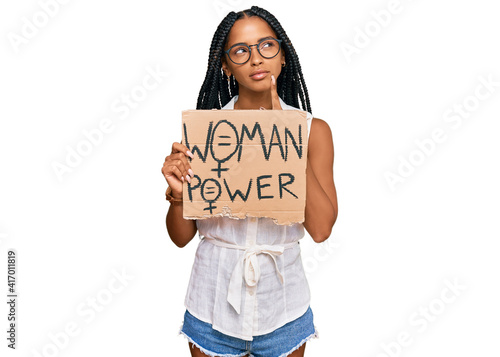 Beautiful hispanic woman holding woman power banner serious face thinking about question with hand on chin, thoughtful about confusing idea