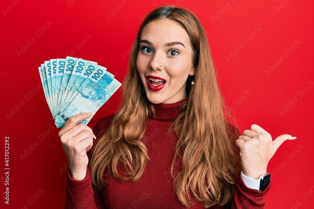 Young blonde woman holding 100 brazilian real banknotes pointing thumb up to the side smiling happy with open mouth Young blonde woman holding 100 brazilian real banknotes pointing thumb up to the side smiling happy with open mouth