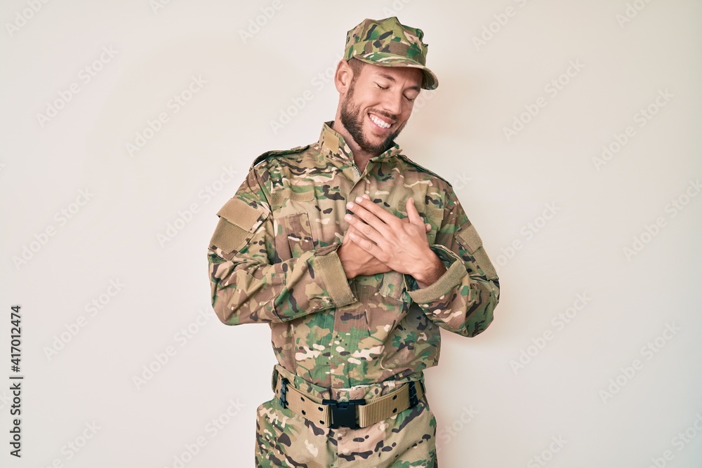 Young caucasian man wearing camouflage army uniform smiling with hands on chest with closed eyes and grateful gesture on face. health concept. Young caucasian man wearing camouflage army uniform smiling with hands on chest with closed eyes and grateful gesture on face. health concept.