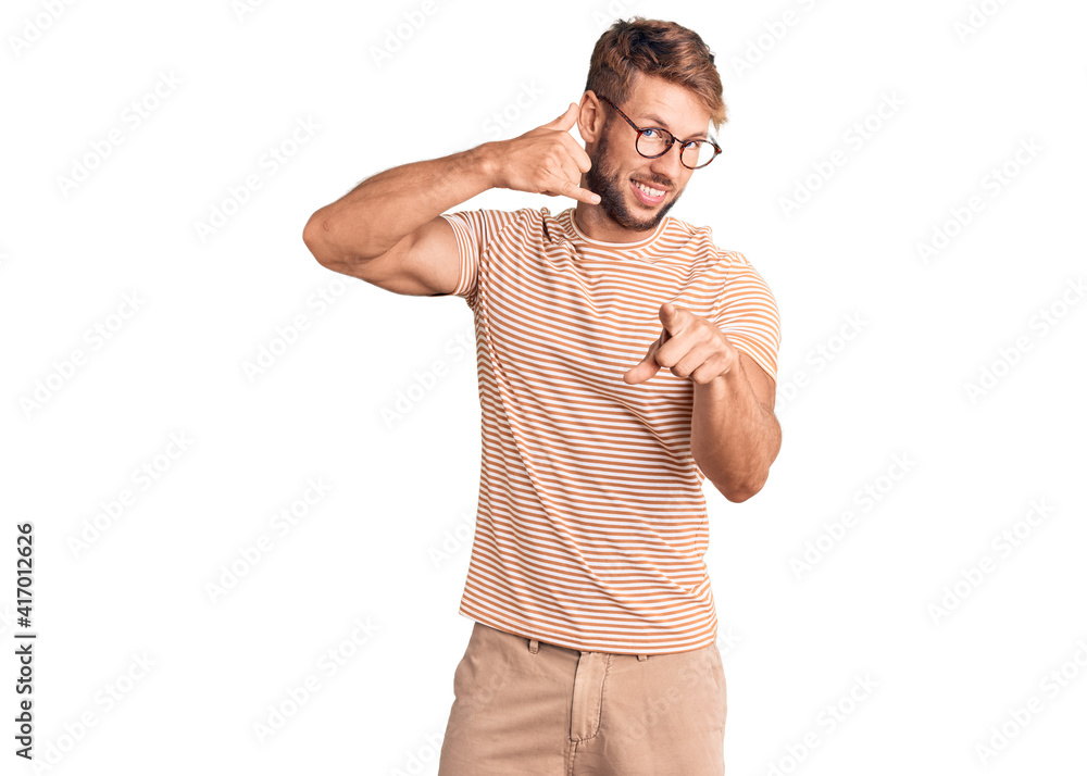 Young caucasian man wearing casual clothes and glasses smiling doing talking on the telephone gesture and pointing to you. call me. Young caucasian man wearing casual clothes and glasses smiling doing talking on the telephone gesture and pointing to you. call me.