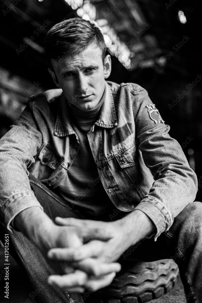 Obraz premium Brutal man in jeans jacket sitting and holding his fingers interlocked. Man portrait