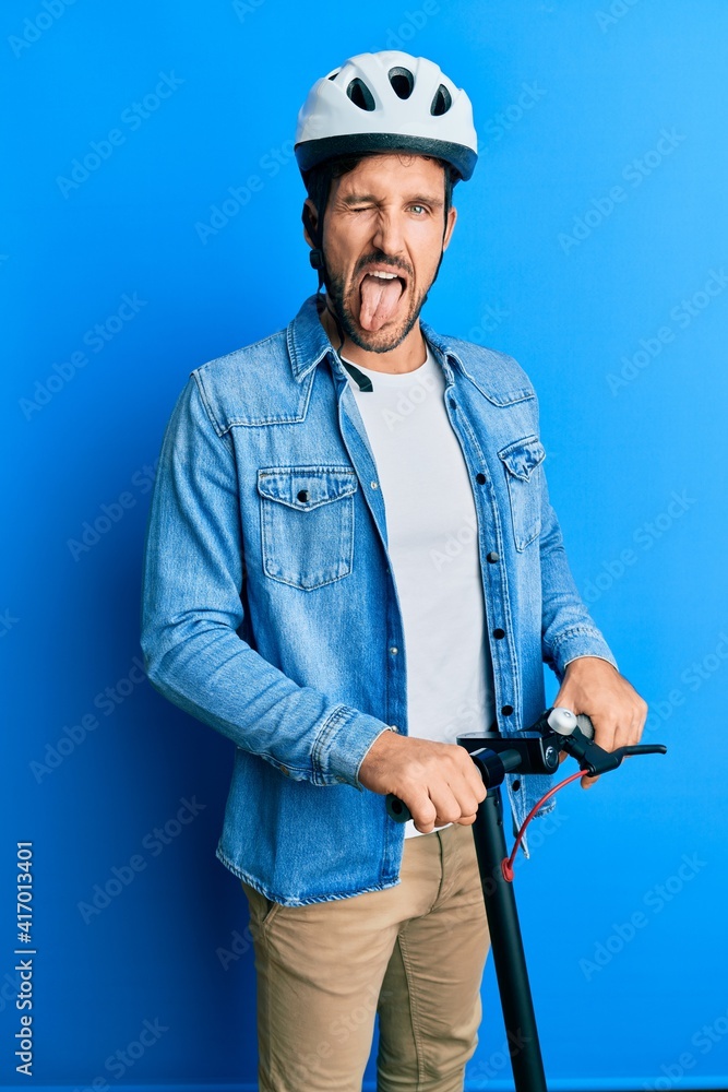 Young handsome man standing on modern scooter wearing helmet sticking tongue out happy with funny expression. Young handsome man standing on modern scooter wearing helmet sticking tongue out happy with funny expression.