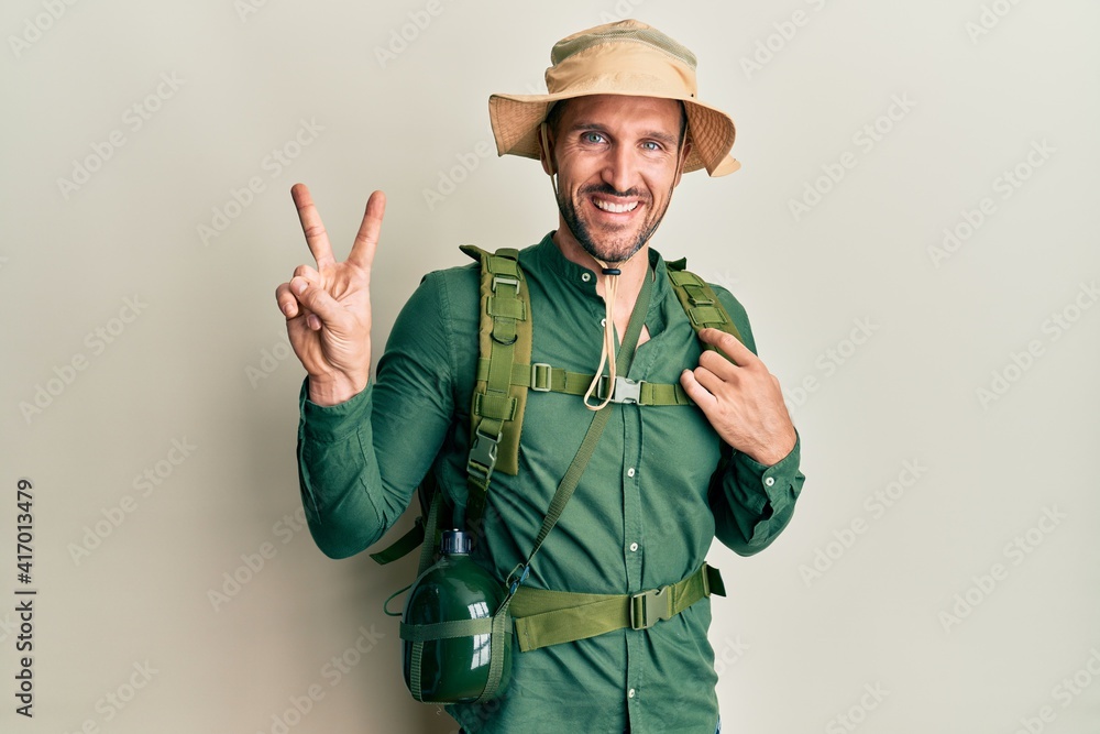 Handsome man with beard wearing explorer hat and backpack smiling looking to the camera showing fingers doing victory sign. number two. Handsome man with beard wearing explorer hat and backpack smiling looking to the camera showing fingers doing victory sign. number two.