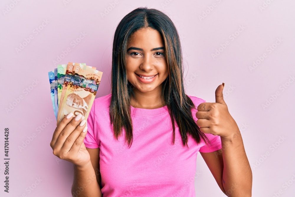 Young latin girl holding canadian dollars smiling happy and positive, thumb up doing excellent and approval sign Young latin girl holding canadian dollars smiling happy and positive, thumb up doing excellent and approval sign