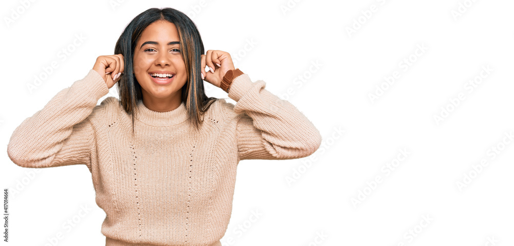 Young latin girl wearing wool winter sweater smiling pulling ears with fingers, funny gesture. audition problem Young latin girl wearing wool winter sweater smiling pulling ears with fingers, funny gesture. audition problem