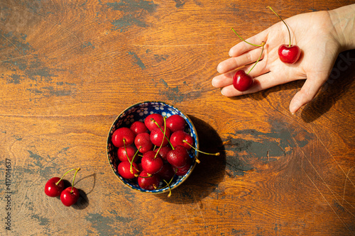 red cherries in bowl and hand on wooden textured background, top view