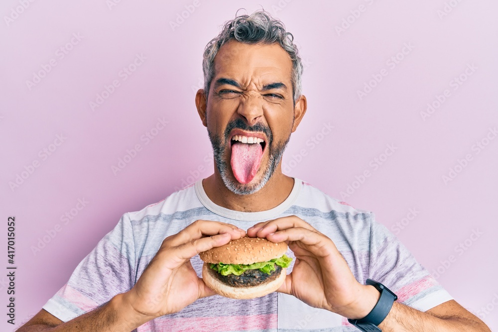 Middle age grey-haired man eating a tasty classic burger sticking tongue out happy with funny expression. Middle age grey-haired man eating a tasty classic burger sticking tongue out happy with funny expression.