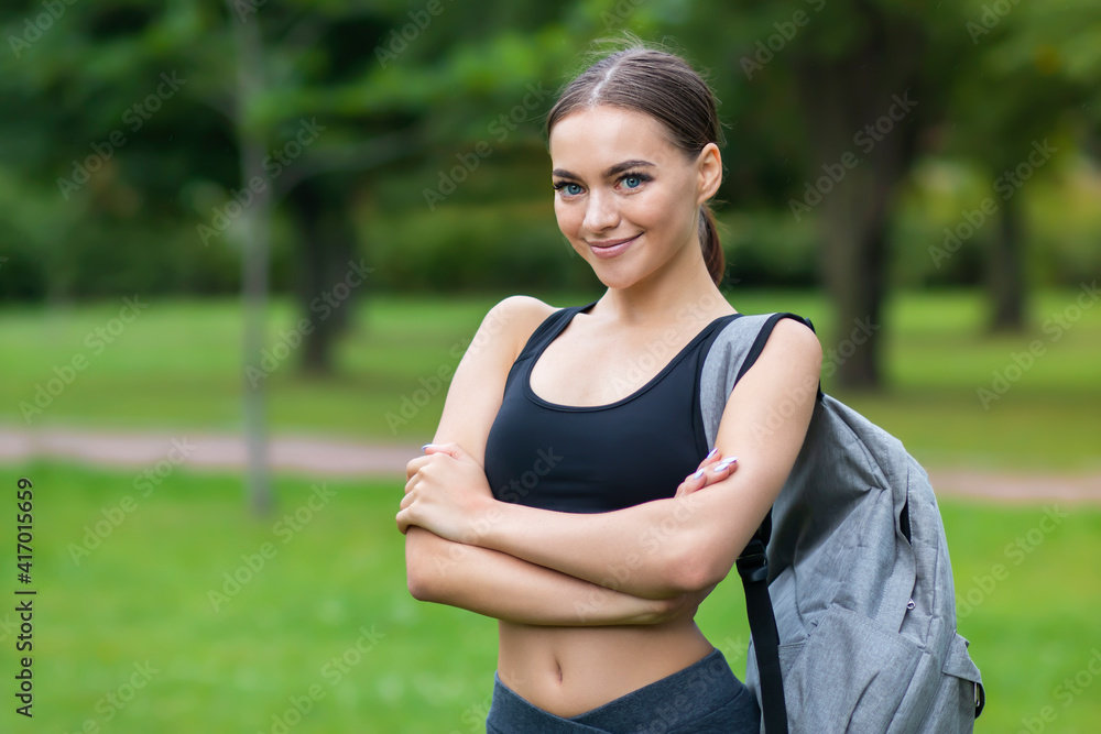 Portrait of happy beautiful positive fit girl, young slim fitness sports woman in sportswear, top outdoors going to training with backpack in park and smiling, looking at camera with her hands crossed