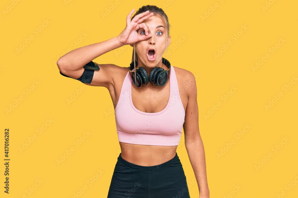 Young blonde woman wearing gym clothes and using headphones doing ok gesture shocked with surprised face, eye looking through fingers. unbelieving expression. Young blonde woman wearing gym clothes and using headphones doing ok gesture shocked with surprised face, eye looking through fingers. unbelieving expression.