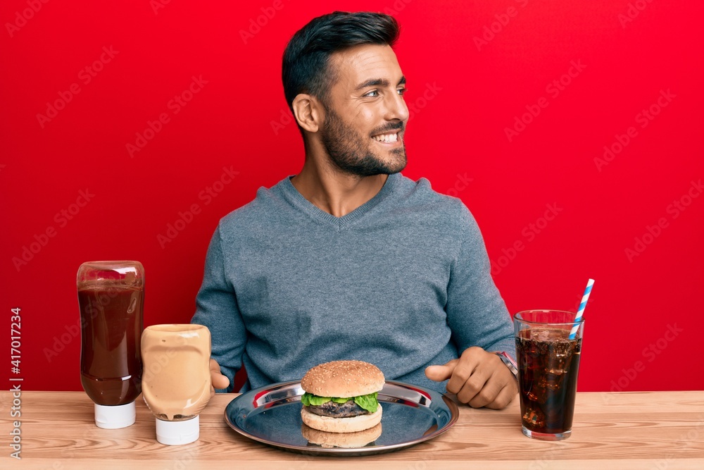 Handsome hispanic man eating a tasty classic burger and soda looking to side, relax profile pose with natural face and confident smile.