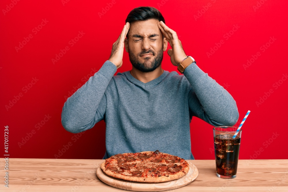 Handsome hispanic man eating tasty pepperoni pizza with hand on head, headache because stress. suffering migraine. Handsome hispanic man eating tasty pepperoni pizza with hand on head, headache because stress. suffering migraine.