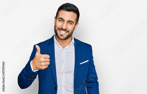 Young hispanic man wearing business jacket doing happy thumbs up gesture with hand. approving expression looking at the camera showing success. photo