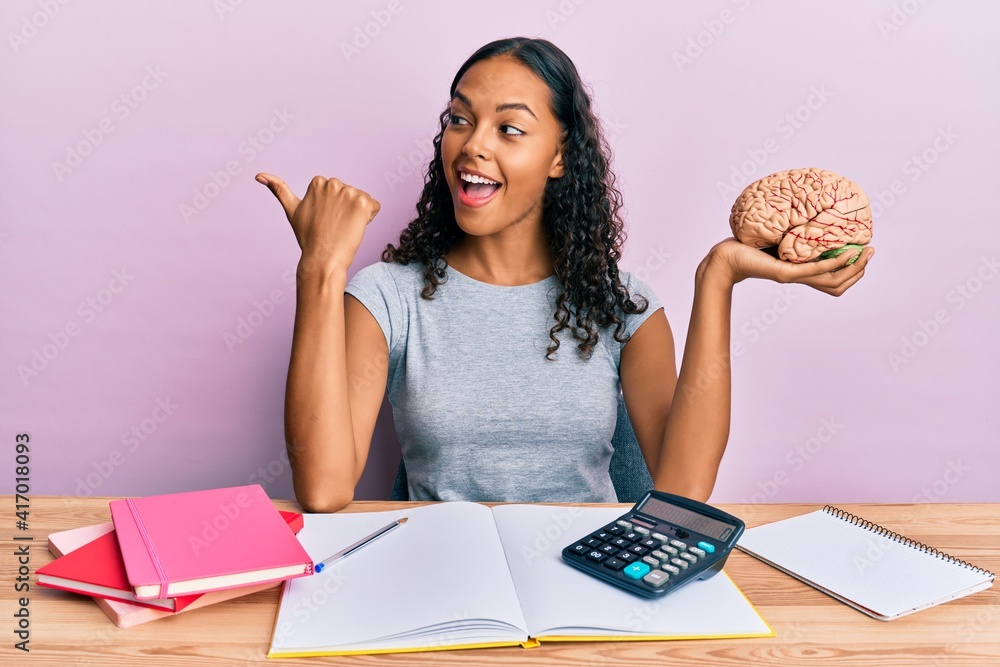 Young african american girl holding brain studying for school pointing thumb up to the side smiling happy with open mouth Young african american girl holding brain studying for school pointing thumb up to the side smiling happy with open mouth